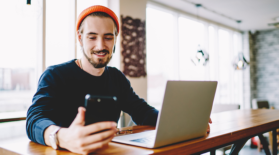 man with a beanie working on a bench with his laptop and phone